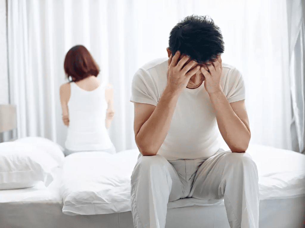 A man looking unhappy about his andropause, with his wife sad in the background.