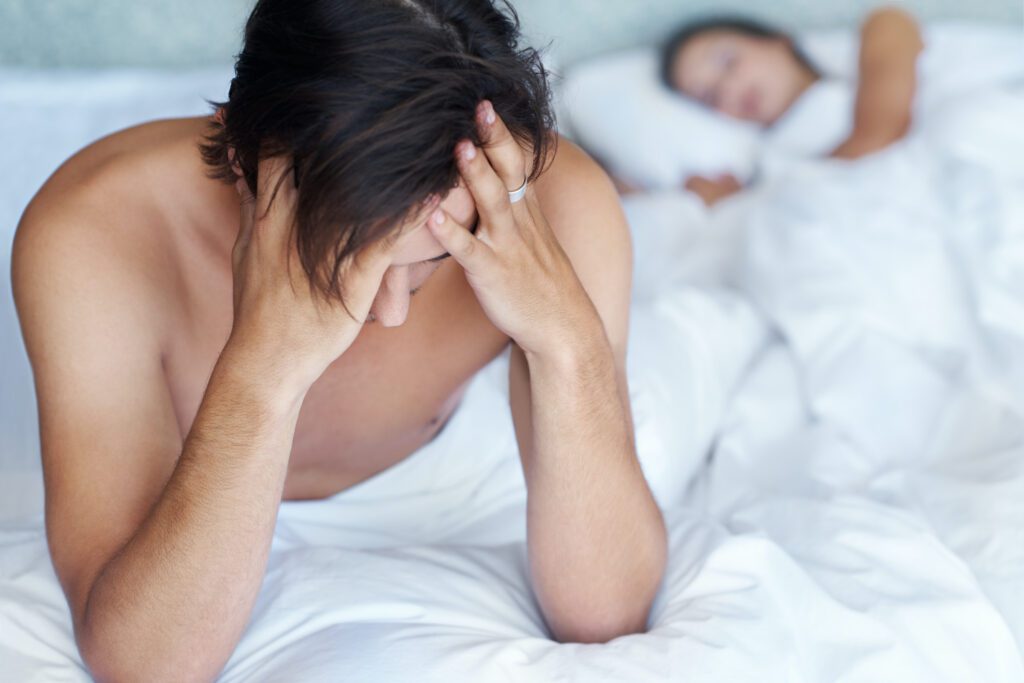 A man sits on the edge of a bed, while someone sleeps in the background.